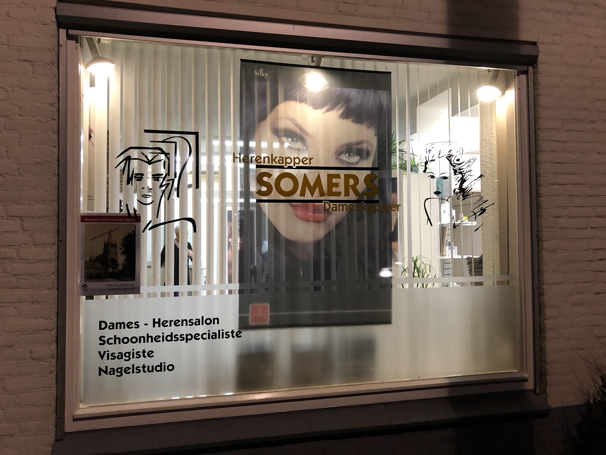 De verlichte etalage van Firma Somers Hairstylist met de diensten op het raam